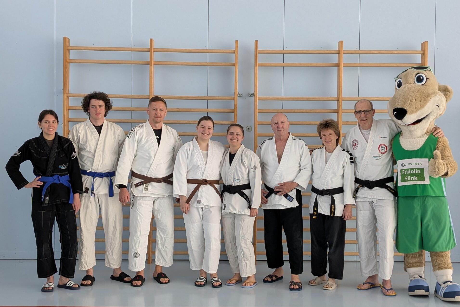 Gruppe von acht Personen in Judokleidung, stehend vor einer Kletterwand, daneben ein Maskottchen in Sportkleidung.