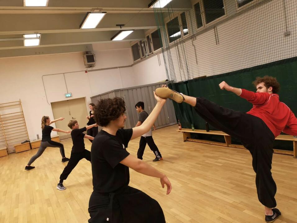 Fünf Personen trainieren in sportlicher Kleidung in einem Raum, wobei eine Person einen Kniestoß ausführt.