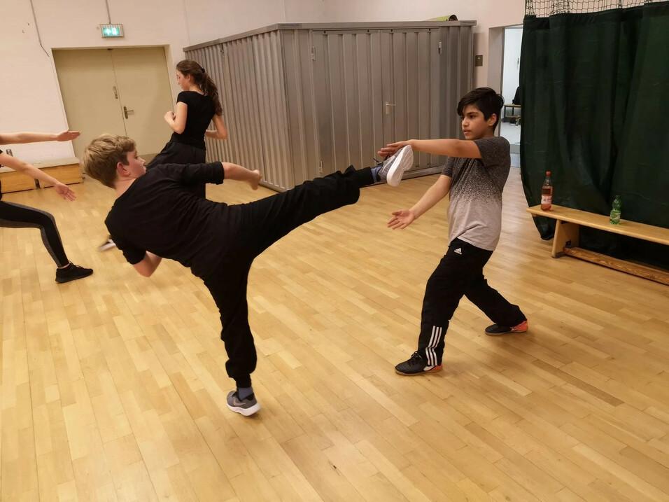 Zwei Kinder üben eine Kampfsporttechnik in einer Turnhalle, während eine weitere Person im Hintergrund steht.