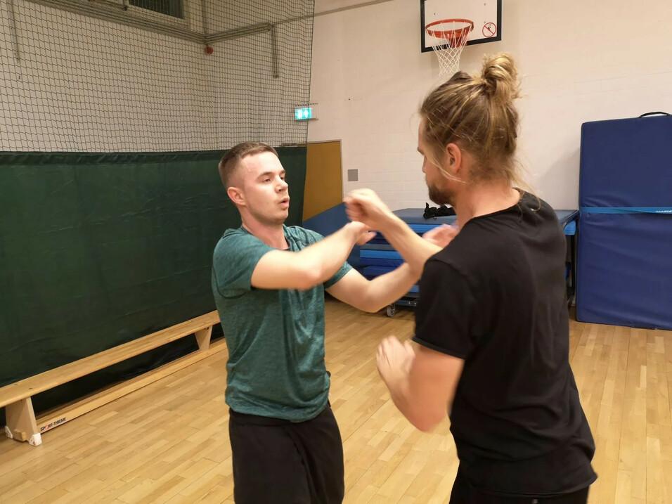 Zwei Personen üben in einer Sporthalle Selbstverteidigungstechniken, während sie sich gegenüberstehen.