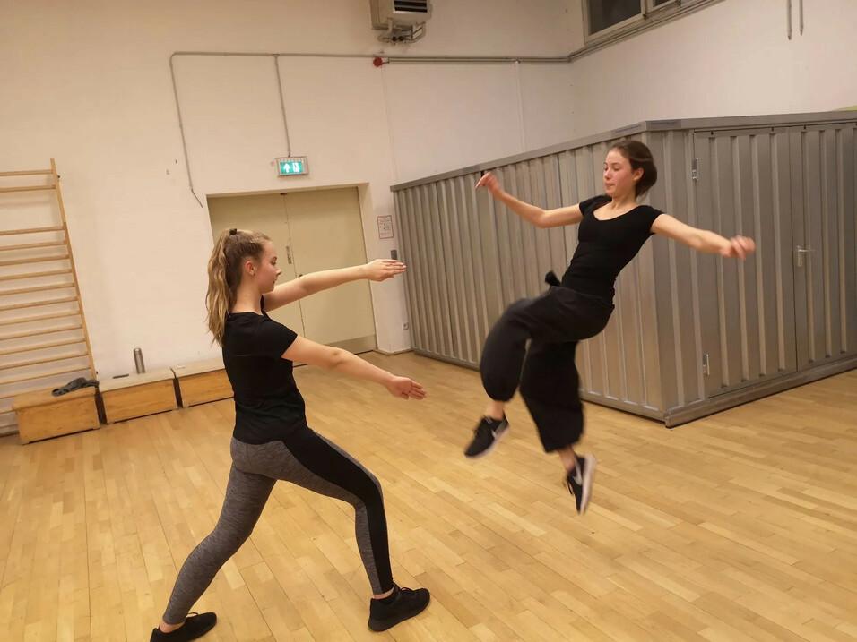 Zwei Tänzerinnen in sportlicher Kleidung, eine springt, während die andere sie auffängt. Hintergrund mit Holzfußboden und Wänden.