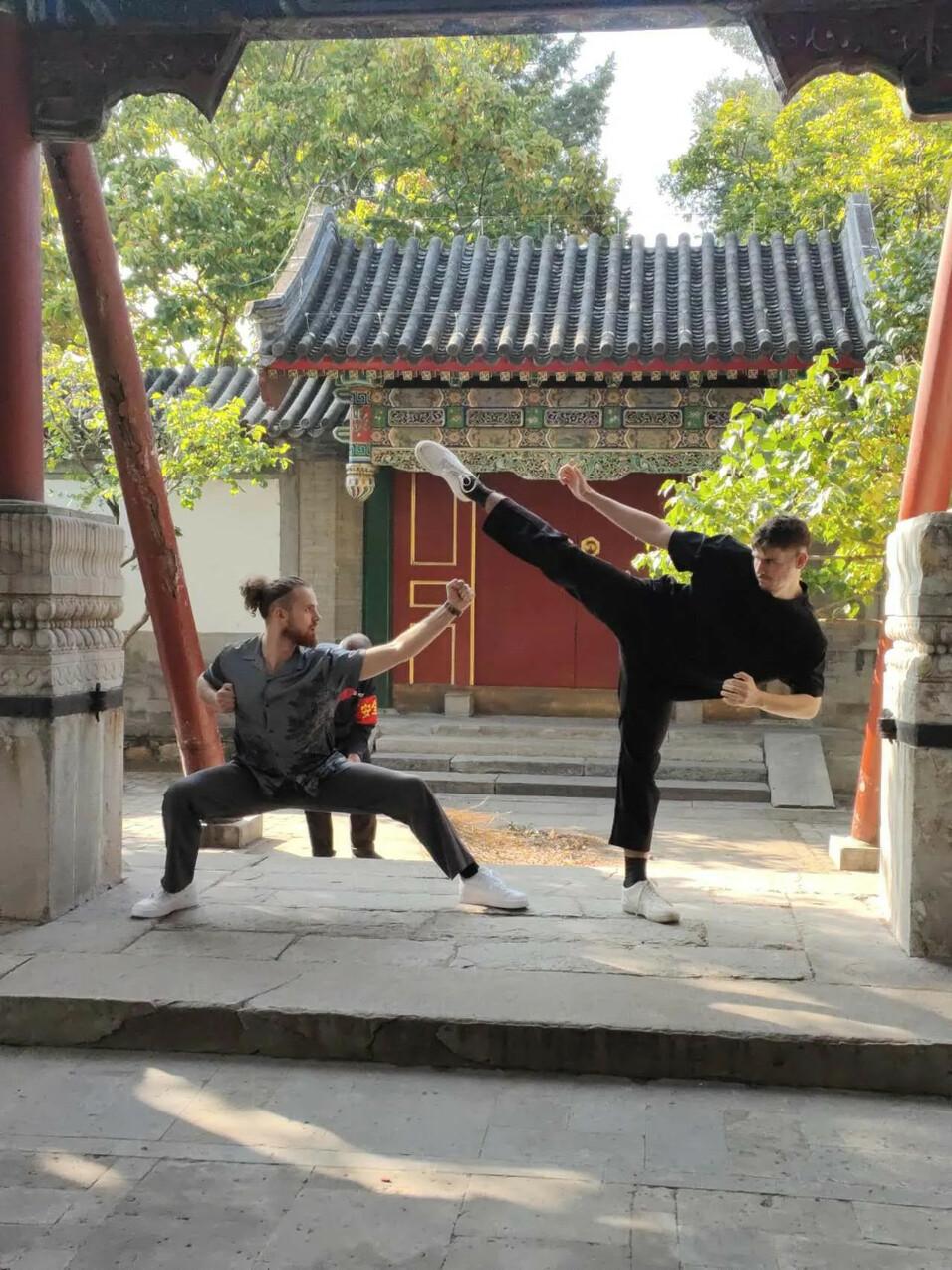 Zwei Männer in Kampfkunstpose vor einem traditionellen chinesischen Gebäude mit grünem Umfeld.