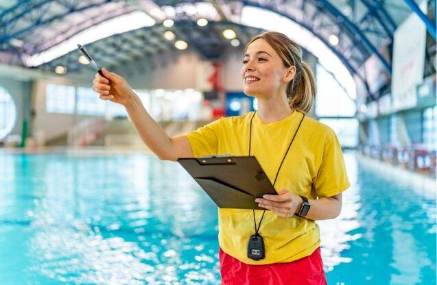 Junge Schwimmtrainerin in gelbem T-Shirt mit Clipboard und Stift am Rand eines Schwimmbeckens.