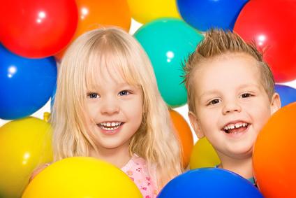 Zwei lächelnde Kinder stehen zwischen bunten Luftballons in Blau, Rot, Gelb und Grün.