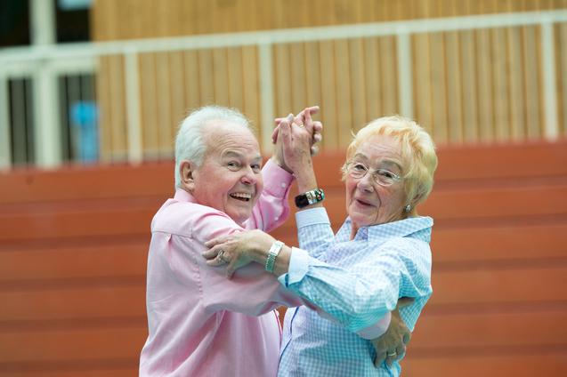 Ein älteres Paar tanzt lächelnd in einer Turnhalle, Kleidung in sanften Farben. Holzpaneele im Hintergrund.