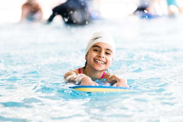Lächelndes Mädchen mit weißer Badekappe schwimmt auf einem Schwimmbrett in einem Pool. Spritzendes Wasser ist sichtbar.